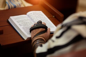 A hand wrapped in tefillin holds an open prayer book with Hebrew text, symbolizing a moment of Jewish prayer and devotion.