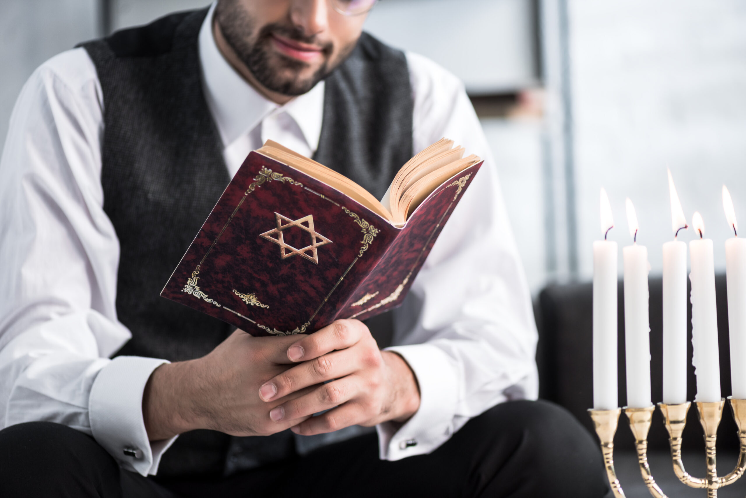 Man in a vest reads a red book with a Star of David on the cover, while candles burn nearby, creating a peaceful, reflective atmosphere.