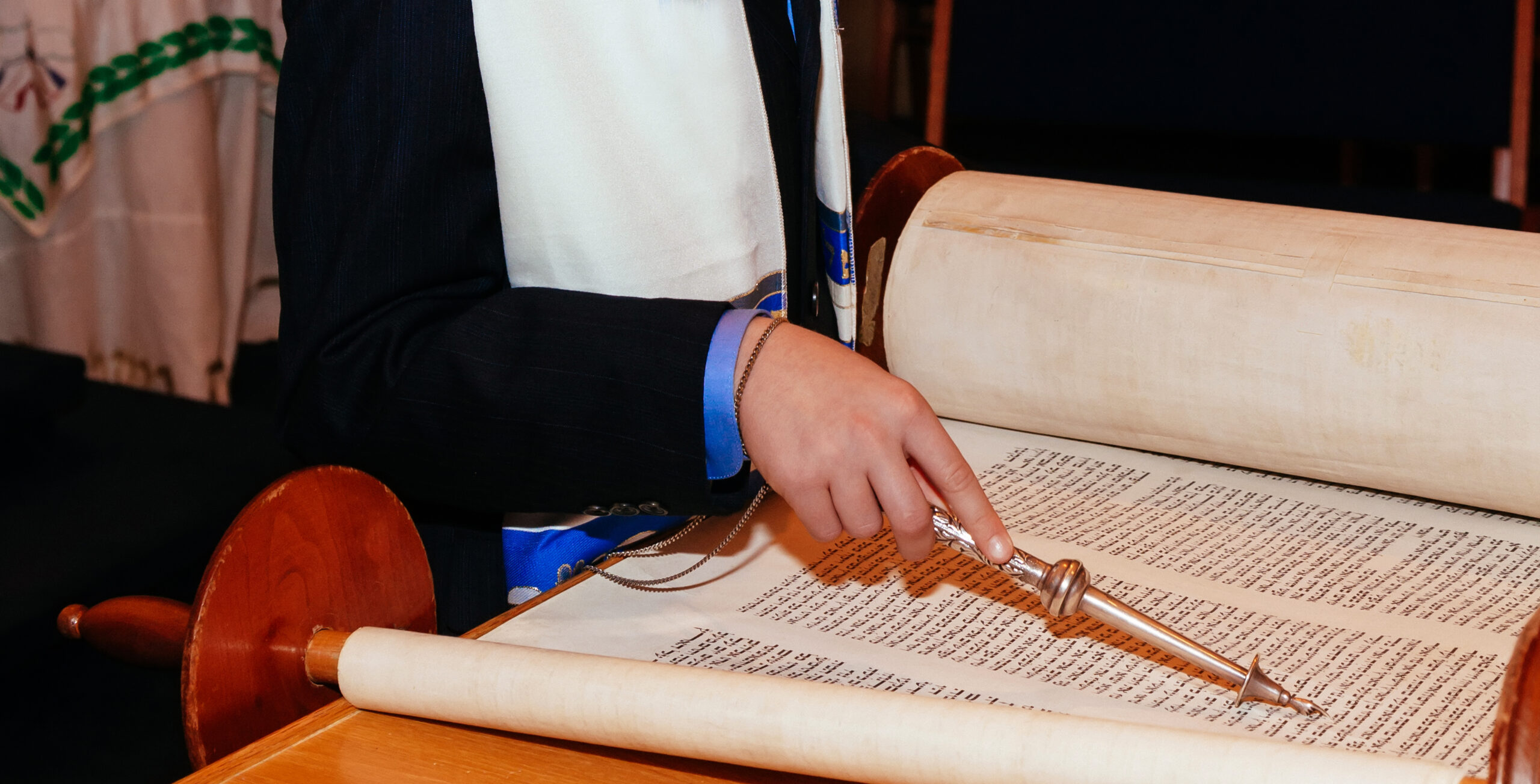 A boy in a formal suit reads from a Torah scroll, using a silver pointer. The setting signifies a Bar Mitzvah or religious ceremony.