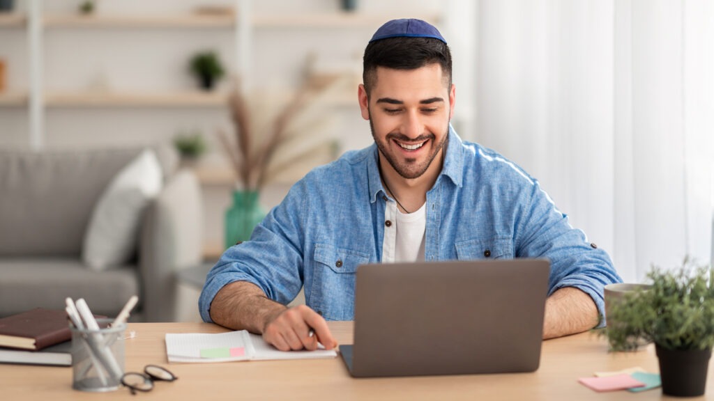 A young man wearing a kippah smiles while working on a laptop. He sits at a wooden desk with notebooks and a plant, indicating a comfortable workspace.