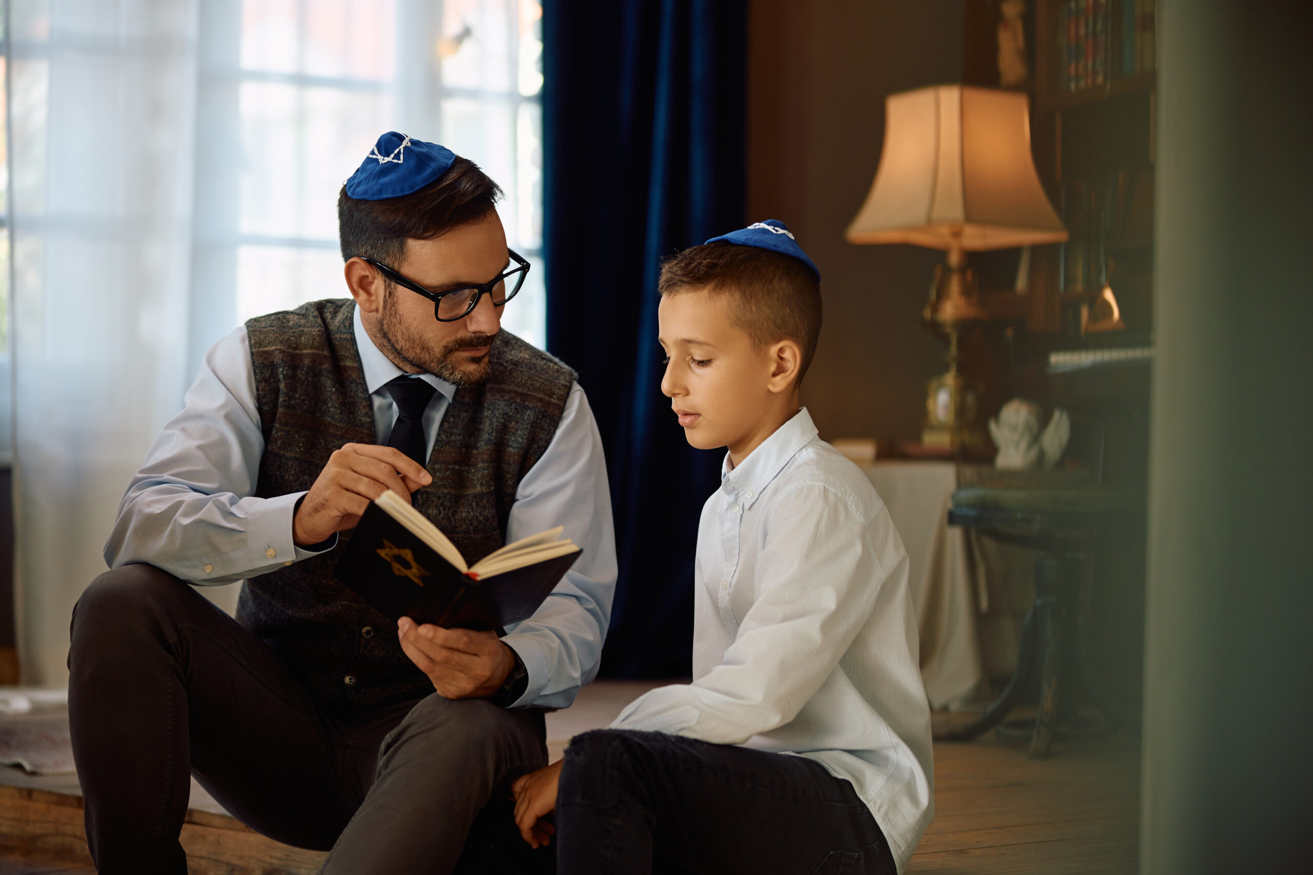 A man and a boy, both wearing blue yarmulkes, engage in a serious discussion while reading a book with a Star of David on the cover.