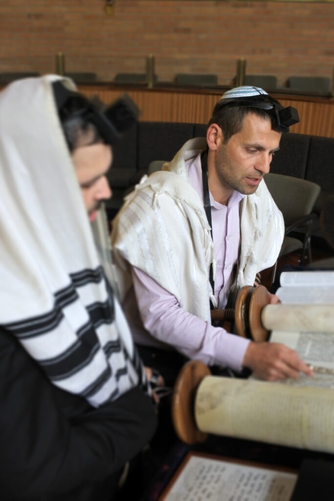 Two men in prayer shawls study a Torah scroll, highlighting a traditional Jewish practice. The setting suggests a synagogue atmosphere.
